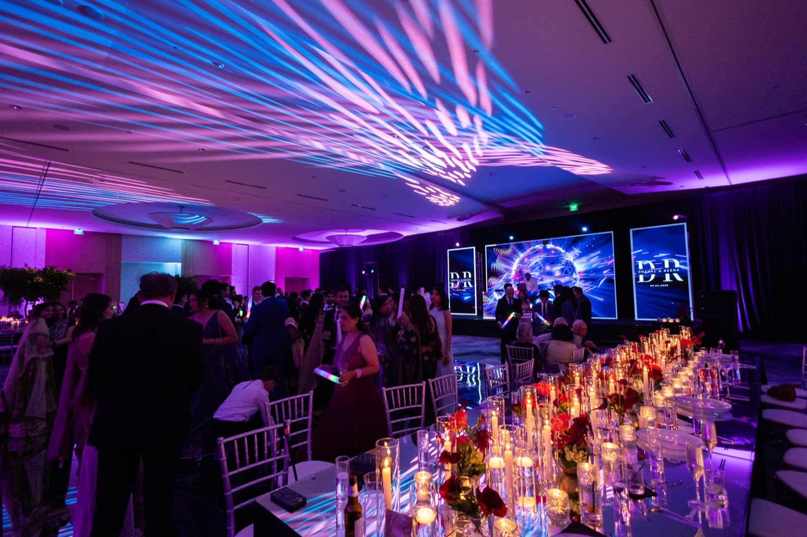 Ballroom during a reception entrance, beam lighting fanned across the ceiling above a long candlelit table set with red roses